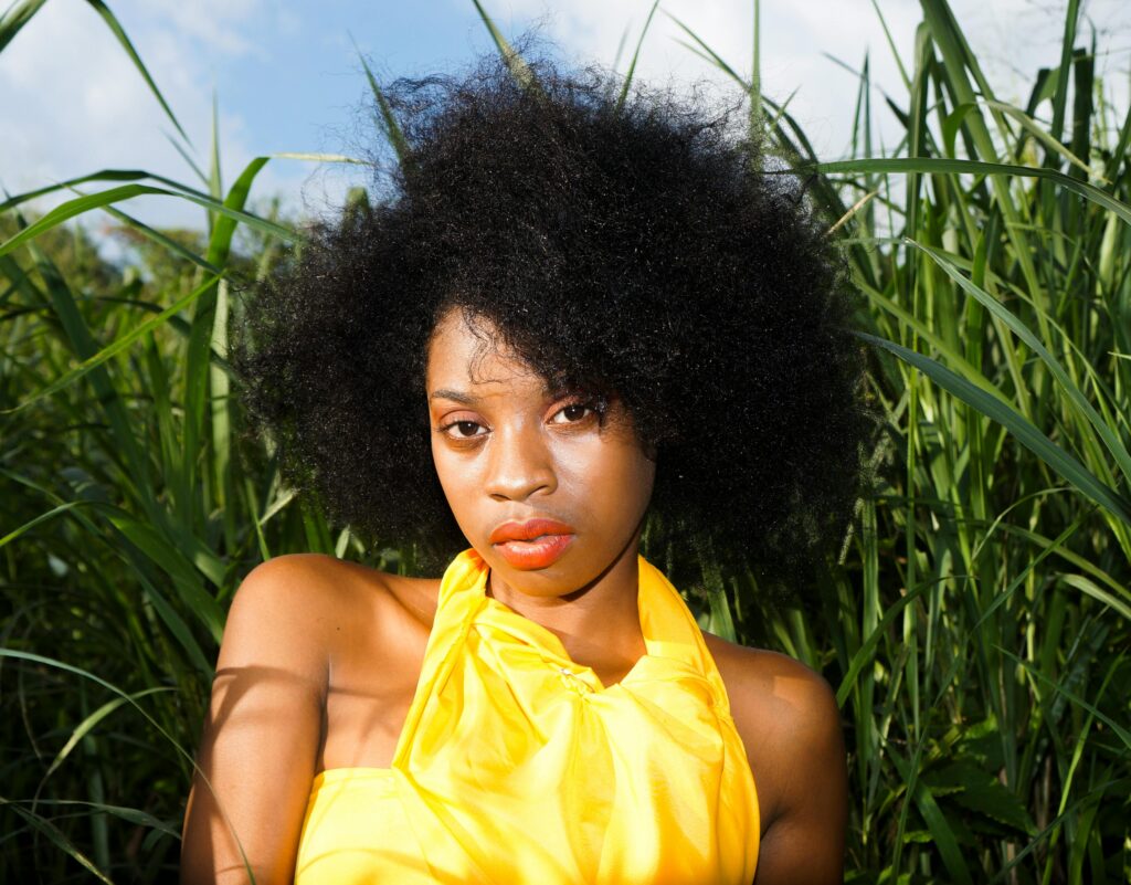 Elegant portrait of a woman with natural afro hair in bright yellow attire amidst tall grass.
